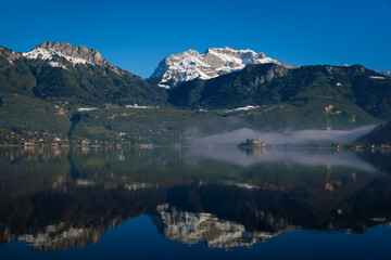 Obraz premium Wide Shot of Lake Annecy Reflecting Mountains and Château de Duingt Surrounded by Fog, France.