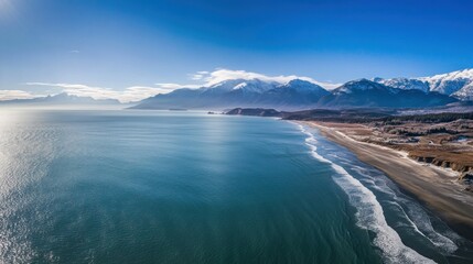 Panoramic aerial view of a pristine beach, ocean, and snow-capped mountains on a sunny day.