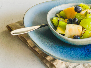 fresh tropical fruit salat desert in blue ceramic bowl on pottery plate and linen napkin on white stone background, colorful mix pineapple, kiwi, blueberries, copy space, high angle view, healthy life