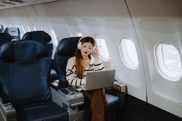 Asian businesswoman wearing headphones, working on her laptop while enjoying music during a flight....