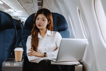 Asian businesswoman is sitting comfortably in airplane seat, working on her laptop with takeaway coffee nearby, enjoying her productive journey