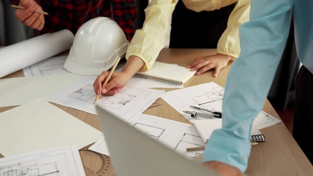 Architect and safety engineer discussion about building blueprint displaying on laptop on table with architectural document and wooden block scatter around at modern office. Closeup. Delineation.