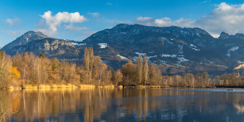 Panorama with Sunset on the Old Rhine with illuminated reeds and bushes on the shore of the lake, sun star and sunset red, impressive colors between autumn and winter, snowy mountains in background