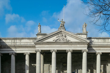 Classical building with tall columns and triangular pediment stands against blue sky. Grand architecture suggest a public building, likely the Bank of Ireland (formerly the Irish Parliament) in Dublin