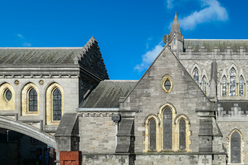 Section of a grey stone building of Christ Church Cathedral in Dublin, Ireland. Blue sky with clouds. Pointed arches frame tall windows with stained glass. Steeply pitched slate roof leads to spire