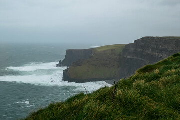 Dramatic coastal scene of Cliffs of Moher in Ireland. Steep cliff drops directly into Atlantic Ocean. Choppy sea with white waves crashing against base of rock formation. Misty and moody atmosphere