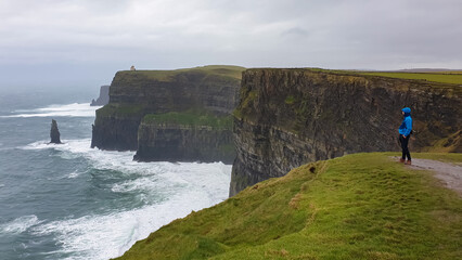 Tourist man with rain protection in dramatic coastal scene of Cliffs of Moher in Ireland. Steep cliff drops into Atlantic Ocean. Choppy sea with waves crashing agains rock formation.Moody atmosphere © Chris