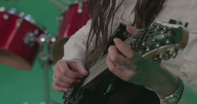 Close-up shot of a woman playing an electric guitar, focusing on her hands on the fretboard. The background is a green screen with red drums slightly out of focus, suggesting a studio setting. 