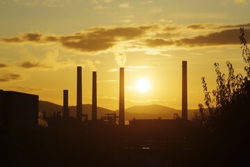 Smoke billowing from industrial chimneys at sunset creates a striking silhouette against the vibrant orange sky, highlighting the stark reality of air pollution and environmental impact