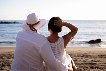 Rear view of bonding senior couple hugging while sitting on the beach face the sunset enjoying vacation and retirement, two elderly people expressing love and tenderness