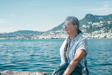Senior white haired woman sitting along lake admiring the landscape - relaxed senior grandmother enjoying peaceful moments in nature, Lake Geneva and mountains in background