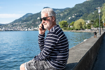 Fototapeta premium Smiling senior white haired man sitting along lake talking on mobile phone - relaxed senior grandfather enjoying peaceful moments in nature, Lake Geneva and mountains in background