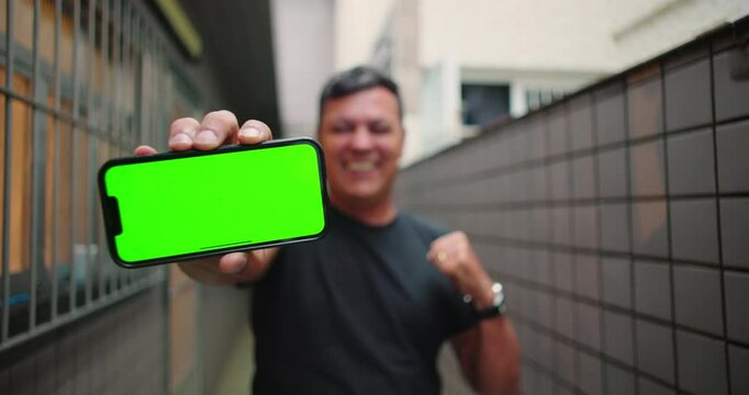 Man celebrating victory with a joyful fist pump while holding smartphone towards the camera, showcasing customizable green screen chroma key, urban corridor background