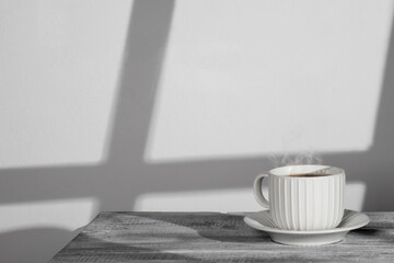 A serene and minimalist photo featuring a white coffee cup on a matching saucer placed on a white wooden table. Steam rises from the freshly brewed coffee, creating a warm and inviting atmosphere. 