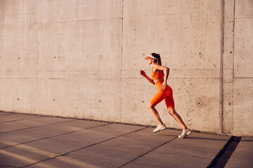 Fototapeta premium Fit woman in orange sportswear running against concrete wall during outdoors exercise