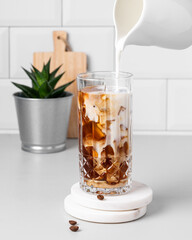 Cold latte coffee in a tall glass with pouring alternative milk from a milk jug on a light table
