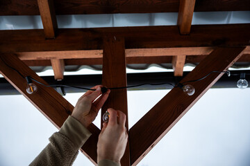 Close up of hands installing LED string light bulbs on a wooden terrace with triangular beams and a corrugated metal roof under an overcast sky.