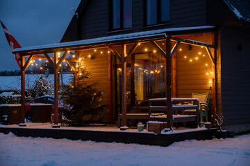 A wooden house terrace with LED string lights, a Christmas tree, outdoor furniture, and snow...