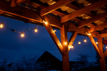 Obraz premium String lights with yellow bulbs hang on a wooden terrace with a corrugated roof. Snow covers the ground and trees under a deep blue evening sky.