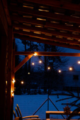 Obraz premium A terrace with warm LED string lights on a wooden pergola, wooden furniture, and a snow covered yard with trees and a building at dusk.