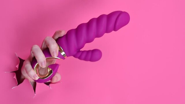 Female hand sticking out with pink sex toy from pink paper background. 