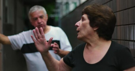 Elderly couple shouting at each other in narrow residential hallway, woman expressing anger while man stands firm, highlighting emotional turmoil in strained relationship - Powered by Adobe