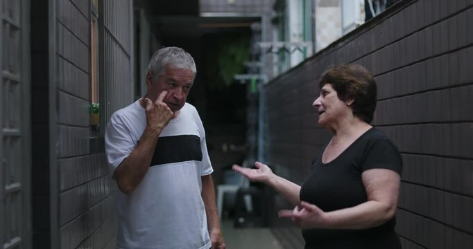 Elderly woman accusing elderly man in pointed gesture during intense argument, while man leans against wall in narrow residential hallway
