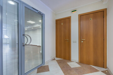 A modern hallway with a glass entrance door and two wooden doors. Natural light illuminates the space, highlighting its clean design and neutral colors.