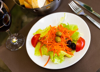There are large pieces of fresh vegetables on plate. Vitamin salad of fresh vegetables, lettuce, olives, tomato, grated carrot is prepared for guests of restaurant