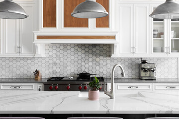 A kitchen detail with metal lights hanging above a large marble island, stainless faucet on a white farmhouse sink, and a marble hexagon tiled backsplash with white cabinets.
