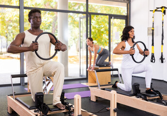 African-american man and hispanic woman kneeling on pilates reformer and doing exercises with rings. Young woman training stretching exercises on pilates chair in background.