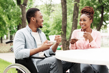 Millennial black guy in wheelchair having date with beautiful woman at outdoor cafe, enjoying coffee and conversation at park. Affectionate African American couple spending time together outside