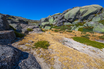 A rugged mountain landscape with large boulders, sparse vegetation, and a distant dam under a clear blue sky