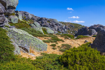 A rugged mountain landscape with large boulders, sparse vegetation, and a distant dam under a clear blue sky