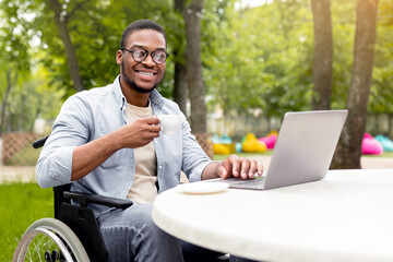 Happy handicapped black man in wheelchair using laptop for online job at cafe in city park, having business meeting on web, outdoors. African American disabled freelancer guy working on remote project