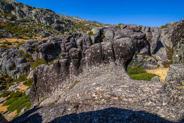 A rugged mountain landscape with large boulders, sparse vegetation, and a distant dam under a clear blue sky