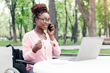 Distance job for people with disability. Happy young black woman in wheelchair working online, using laptop, speaking on smartphone and enjoying coffee at outdoor cafe, copy space