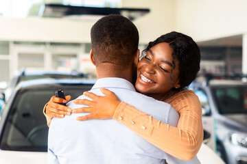 Grateful Black Wife Embracing Husband After Buying Car In Dealership Showroom, Happy African American Woman Thankful For Purchasing New Automobile, Cheerful Lady Holding Keys And Smiling, Closeup