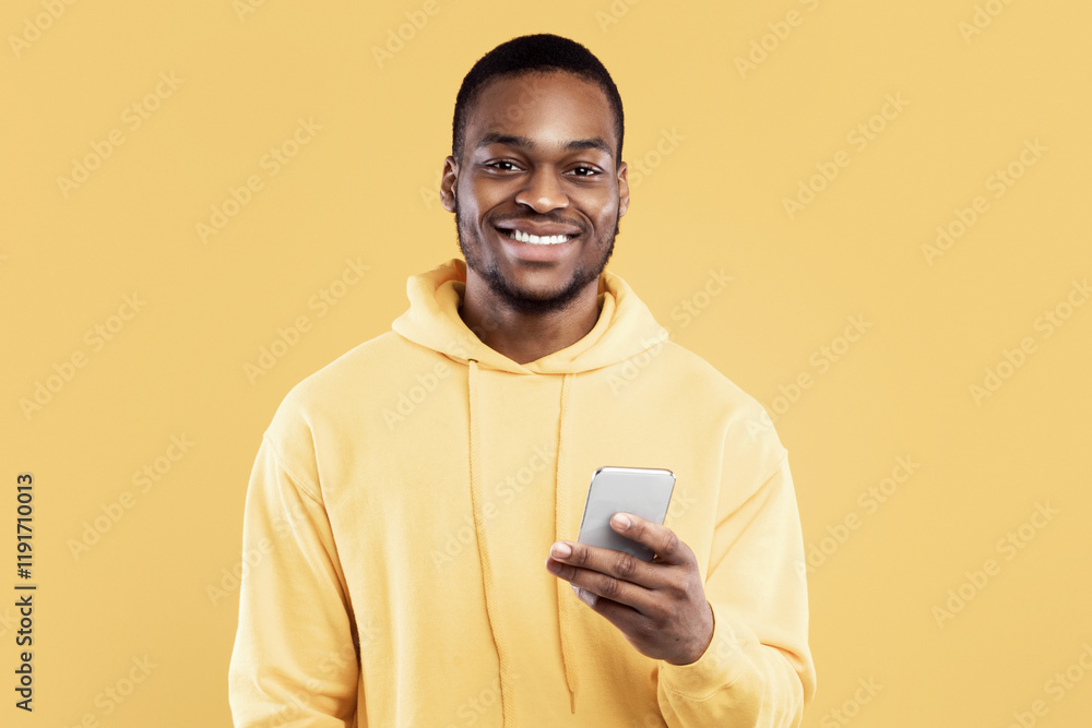 Poster happy black millennial man using mobile phone smiling looking at camera standing over yellow studio 