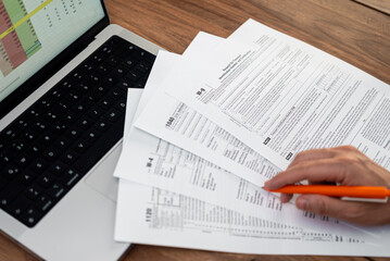 Fototapeta premium Close-up of accountant's hands reviewing tax forms with orange pen and laptop on wooden table. Financial planning and tax preparation concept 
