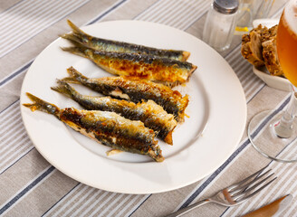 Golden-brown grilled sardines served on white plate, paired with crusty bread and refreshing glass of beer