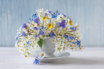 Still life with a bouquet of spring white flowers of bird cherry, blue pansies, scilla and brunnera in a cup on a table