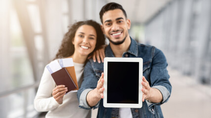 Arab Couple Demonstrating Digital Tablet With Black Screen While Standing In Airport With Passports And Tickets In Hands, Young Spouses Showing Copy Space For Your Design Or Advertisement, Mockup