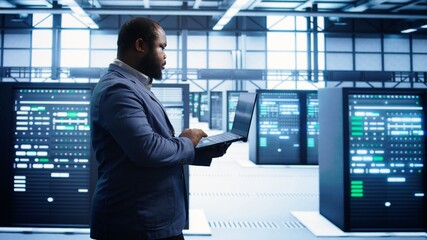 African american man walking in high tech facility, using laptop to patch network issues leading to...