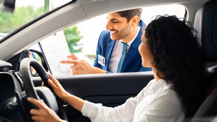 Young long-haired brunette woman sitting inside brand new comfy car, having conversation with...