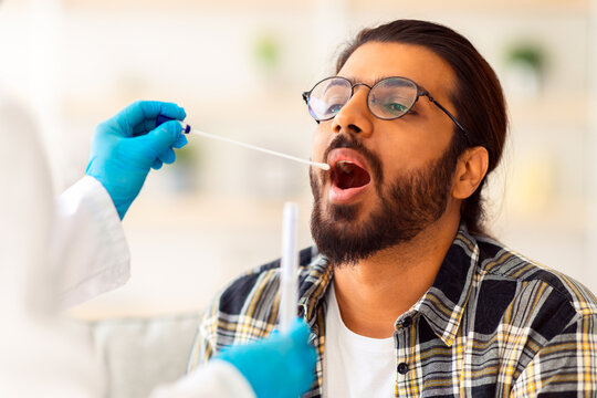 Indian guy getting coronavirus swab test from throat at home, unrecognizable nurse in protective medical costume making covid-19 swab test for sick patient young man at home, closeup - Powered by Adobe