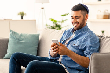 Obraz premium Mobile Communication Concept. Portrait of happy young jewish man using modern smartphone device sitting on comfy sofa at his apartment. Cheerful guy typing sms message, browsing social network