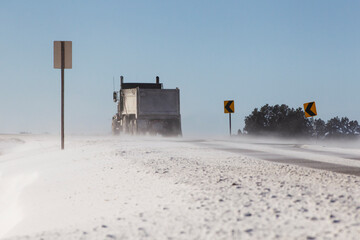 A lone dump truck drives along snow-covered roads with directional arrow signs against a clear blue...