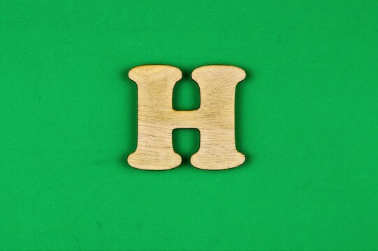 A close-up of a light brown wooden letter "H" positioned against a bright, vibrant green surface.
