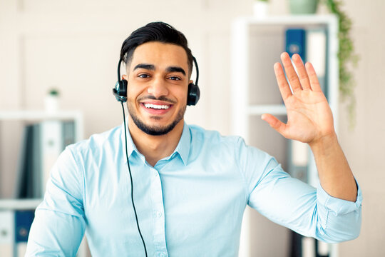 Remote communication concept. Happy Arab guy with headset having video call at office, waving at webcam. Head shot of young man using computer for business meeting or web conference - Powered by Adobe
